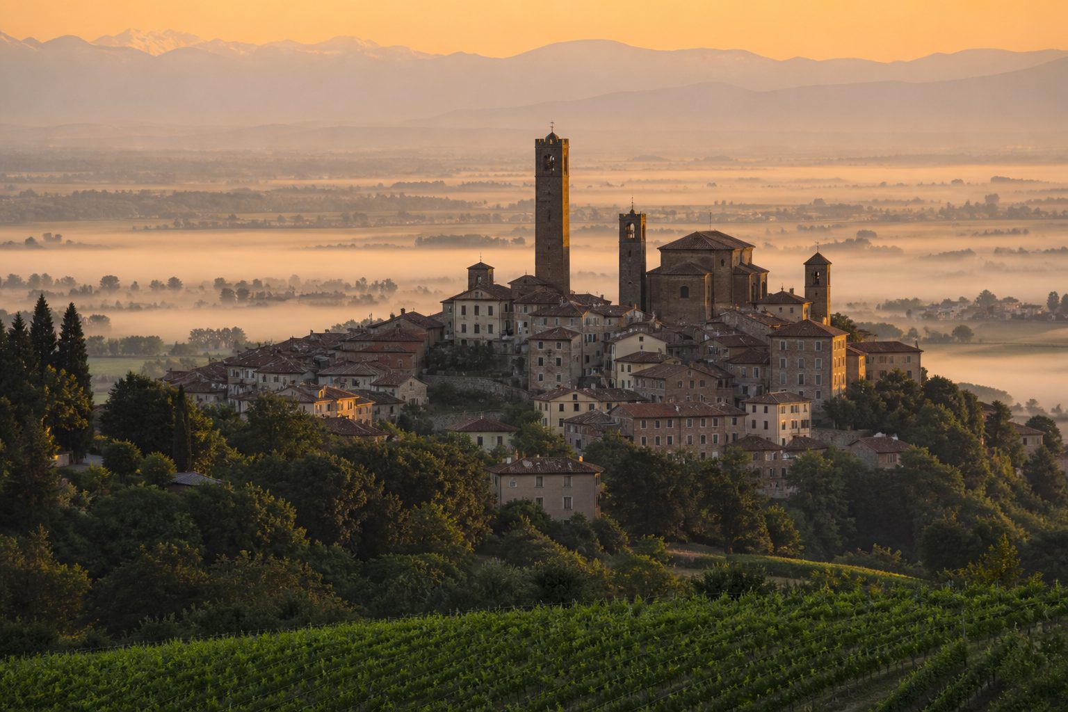 Cittadina medievale compatta vista da una collina all'alba, con torri in mattoni sopra i tetti e foschia sulla pianura
