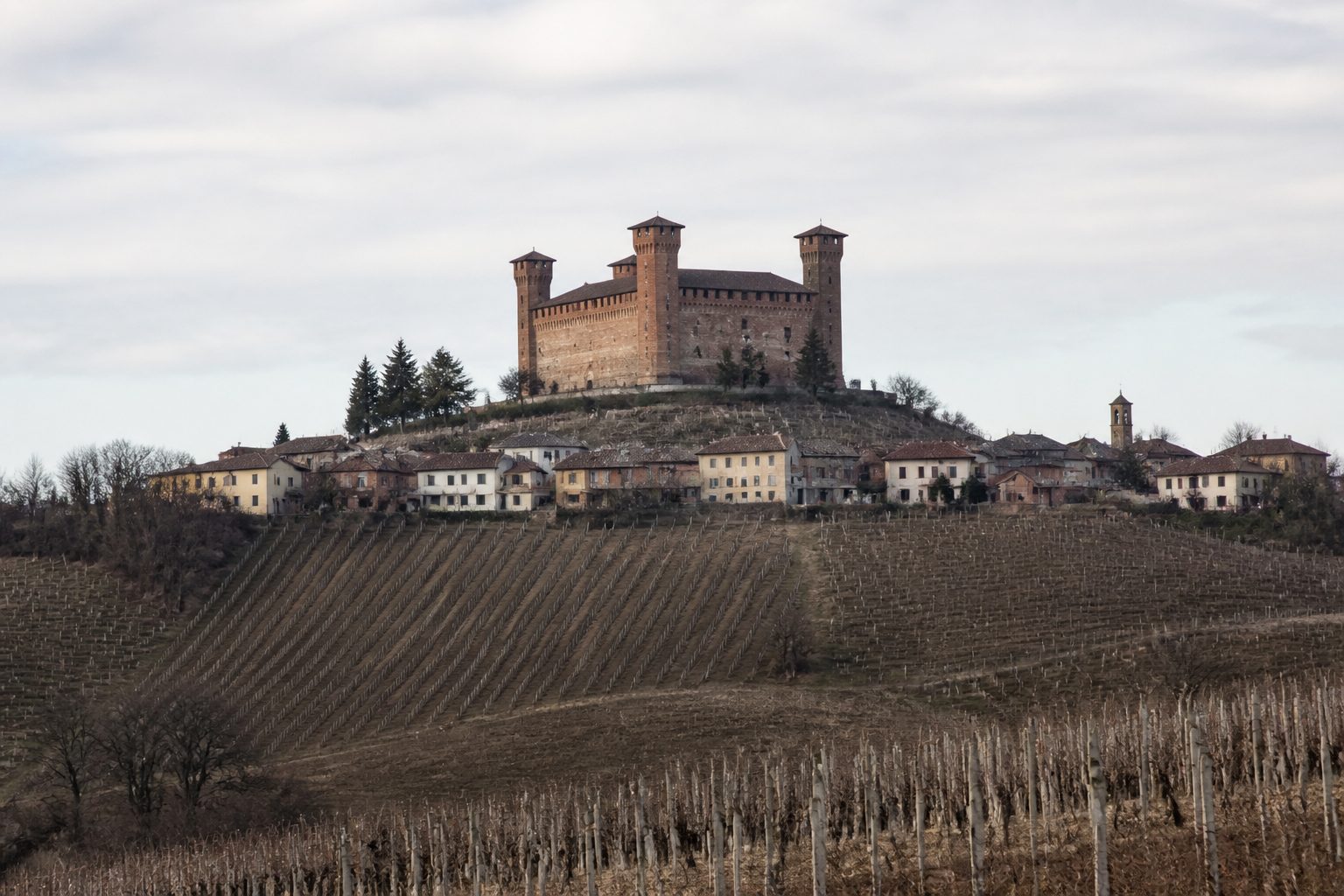 Castello medievale in mattoni con torri angolari sopra un paese, vigneti spogli intorno e luce invernale