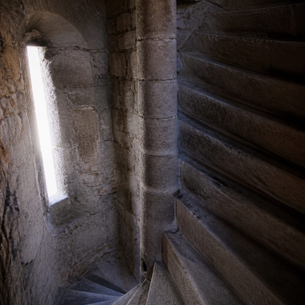 Scala a chiocciola in pietra all'interno di una torre, gradini consumati e feritoia che filtra la luce