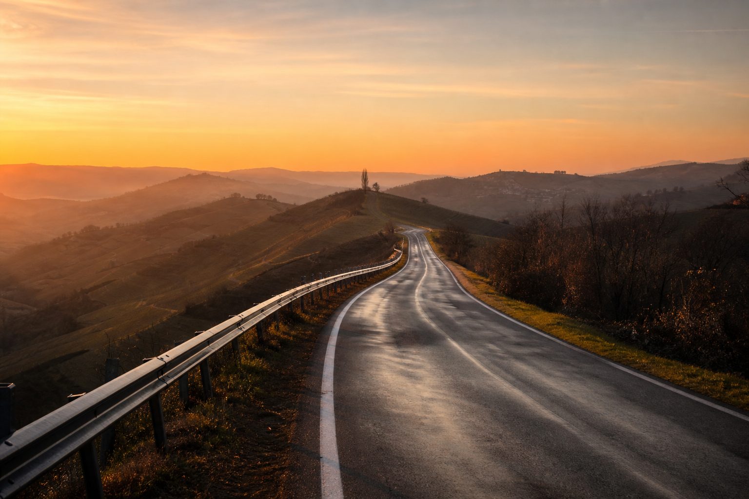 Strada deserta che corre su un crinale alle prime luci, asfalto ancora bagnato e colline sovrapposte